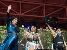 Three drag performers in colorful outfits wave on an outdoor stage under red rafters and lights, smiling at the crowd.