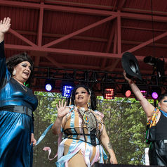 Three drag performers in colorful outfits wave on an outdoor stage under red rafters and lights, smiling at the crowd.