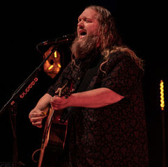 Long-haired man plays guitar and sings passionately on stage, under warm red lighting. Dark background with glowing orange lights.