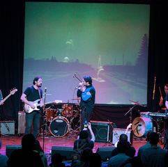 Band performing on stage, with a guitarist, singer, drummer, and keyboardist. A landscape projection in the background. Audience watching.