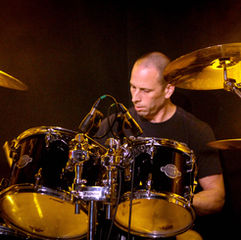 Drummer plays drums on stage, focused expression. Black drum set with "Sonor Ascent" logo. Warm lighting, dark background.