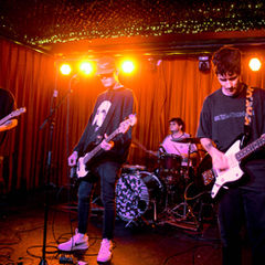 A band performs on stage with guitars and drums. Orange lighting, red curtains, and vibrant energy create a lively concert atmosphere.