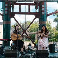 Two women play acoustic guitars and sing on an outdoor stage with microphones and a keyboard, framed by trees and blue sky.