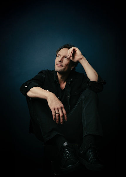 Thoughtful man in black clothes sits against a dark blue studio background, resting his head on his hand.