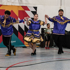 Three folk dancers in blue and gold costumes perform on a stage before a colorful geometric mural, with a child watching.