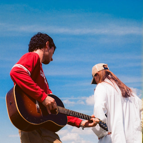 Man in red sweater playing a guitar outdoors, beside a person in a white shirt and cap, against a bright blue sky background.