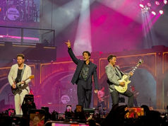 The Jonas Brothers perform energetically on stage amid colorful lights. Two hold a guitar; crowd captures the moment with phones.