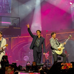 The Jonas Brothers perform energetically on stage amid colorful lights. Two hold a guitar; crowd captures the moment with phones.