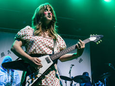 Woman in a red polka-dot dress plays electric guitar under green stage lights, with a blurred band backdrop.