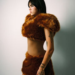 Person with tattoos in a brown furry outfit stands against a plain white backdrop, looking determined. Neutral expression.