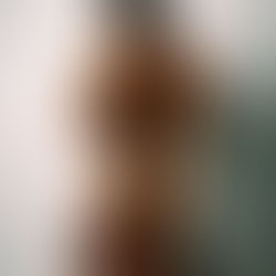 Person with tattoos in a brown furry outfit stands against a plain white backdrop, looking determined. Neutral expression.