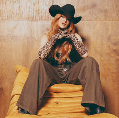 Savannah Jade in a cowboy hat and leopard print gloves sits confidently on a yellow couch against a wood background, wearing brown pants.