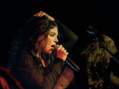 Female singer on a dark stage sings into a microphone, hand in hair, with a blurred guitarist behind her.