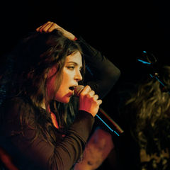 Female singer on a dark stage sings into a microphone, hand in hair, with a blurred guitarist behind her.