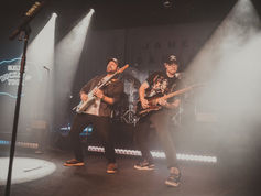 Two guitarists perform energetically on a foggy stage, lit by spotlights. A neon sign and partial "James Band" text are visible in the background.