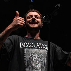 Man on stage giving thumbs up, wearing "IMMOLATION" t-shirt. Dark background, microphone visible. Joyful expression.