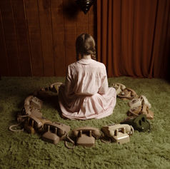 Girl in a pink dress sits on a green shag rug, surrounded by a circle of old rotary phones in a dim room.
