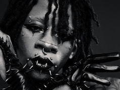 Close-up of a person with black lipstick, facial piercings, and spiked gloves. Dramatic makeup and intense expression in monochrome.