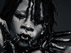 Close-up of a person with black lipstick, facial piercings, and spiked gloves. Dramatic makeup and intense expression in monochrome.