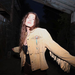 Woman in a fringed tan jacket poses with arms outstretched in a dimly lit rustic setting. Confident expression, night scene.