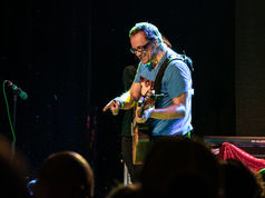 Musician in blue shirt playing guitar and smiling on stage, with a microphone nearby and dim background lighting. Enthusiastic vibe.