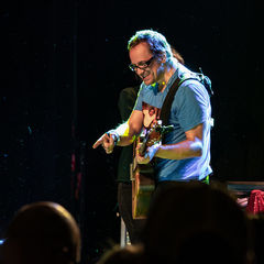 Musician in blue shirt playing guitar and smiling on stage, with a microphone nearby and dim background lighting. Enthusiastic vibe.