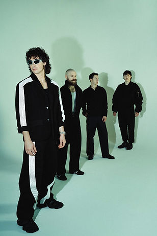 Four men in black outfits stand in a green-lit room. One wears sunglasses with striped pants. The mood is calm and mysterious.