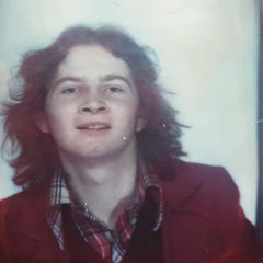 Young person with medium-length hair smiles, wearing a red jacket and plaid shirt. Neutral background with a nostalgic, vintage feel.