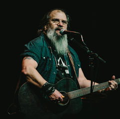 Bearded singer with glasses plays acoustic guitar and sings into a mic on a dark stage, focused and intimate.