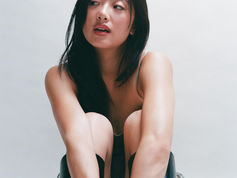 Woman sitting with knees up, wearing black boots, against a light background. She looks thoughtful, with long hair framing her face.