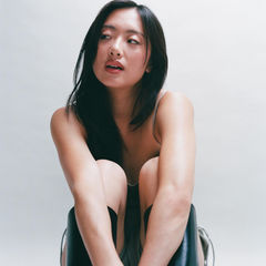 Woman sitting with knees up, wearing black boots, against a light background. She looks thoughtful, with long hair framing her face.