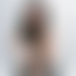 Woman sitting with knees up, wearing black boots, against a light background. She looks thoughtful, with long hair framing her face.