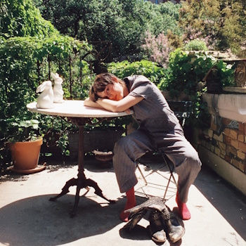 Person in gray outfit resting head on a round table on a sunlit patio, surrounded by greenery. Red socks and a ceramic rabbit are visible.