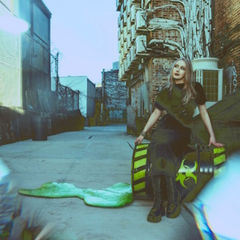 Woman in black sits on a toxic barrel with green slime in an urban alley. Brick wall background, graffiti, and industrial elements visible.