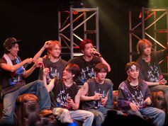 The seven members of Travis Japan on stage, wearing graphic shirts and jeans, with one singing into a microphone. Bright lights and a lively mood.