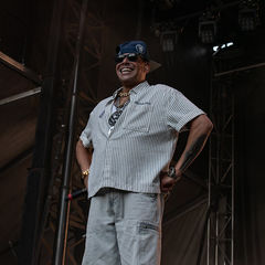 Smiling man on a concert stage, hands on hips, wearing a striped shirt, gray pants, blue cap, and sunglasses amid dark rigging.