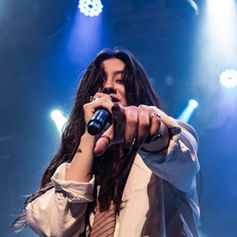 Performer (Jade LeMac) on stage points forward, holding a mic. Blue and purple lights illuminate the background, creating a dynamic, energetic atmosphere.
