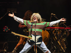 Blond singer-guitarist performs on a dark stage with arms outstretched, singing into a mic; band gear and nord wave2 behind.