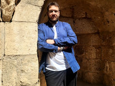 Man in blue shirt leans against stone archway, arms crossed. Sunlight illuminates the scene, creating a relaxed, contemplative mood.