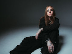 Person with long hair and tattoos sits on the floor in a dimly lit room, wearing black clothes. The mood is contemplative.