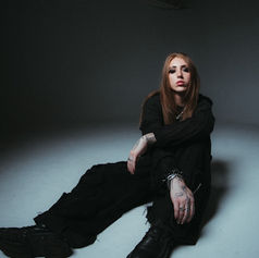 Person with long hair and tattoos sits on the floor in a dimly lit room, wearing black clothes. The mood is contemplative.