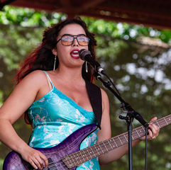Woman singing into a microphone while playing a purple bass on an outdoor stage, with a rainbow flag and green trees behind.