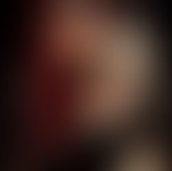 Person with red hair and intricate makeup holds skulls, wearing black gothic attire. Dark background enhances spooky, mysterious mood.