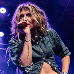 Singer in a denim shirt performing passionately under purple stage lights, holding a red microphone. Tattoo visible on arm.