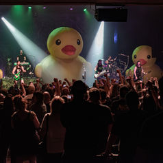 Metal band Alestorm performs on stage with three giant inflatable yellow ducks as the crowd cheers and raises fists under bright spotlights.