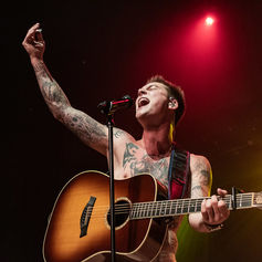 Tattooed singer, Cameron Whitcomb, passionately performs on stage with an acoustic guitar under red and yellow lights. Emotive and dynamic atmosphere.