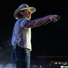 Tattooed singer in a cowboy hat points a microphone on a dark, smoky stage under concert lights.