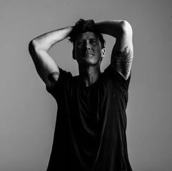 A person in a black T-shirt poses with hands on head against a gray background. They have tattoos on both arms, conveying a relaxed mood.
