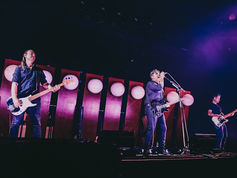 Three musicians perform on a purple-lit stage, singer at microphone center with guitars on either side and round lights behind.