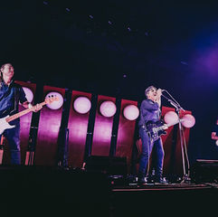 Three musicians perform on a purple-lit stage, singer at microphone center with guitars on either side and round lights behind.